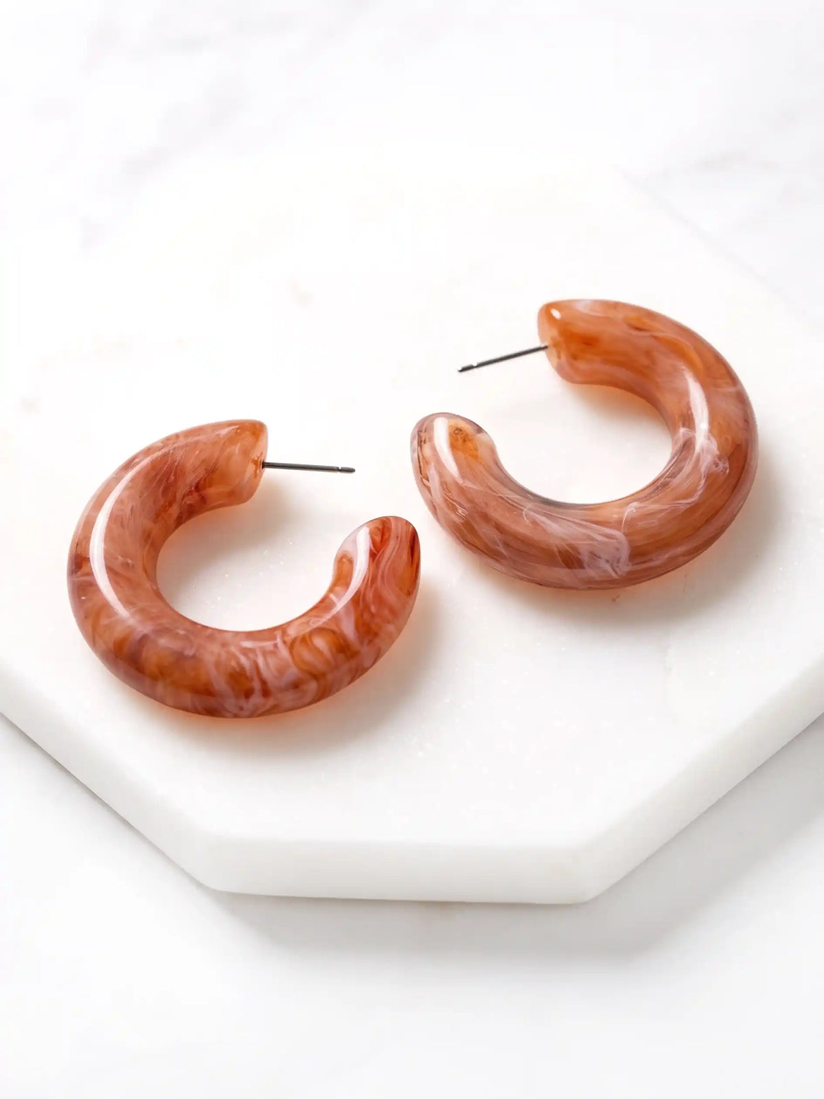 A pair of chunky, open C-shaped resin hoop earrings featuring a swirled brown and white marbled pattern, resting flat on a white marble coaster.