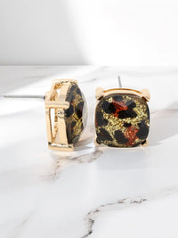 A pair of gold-tone stud earrings with cushion-cut black and gold marbled resin centers, displayed on a white marble surface.