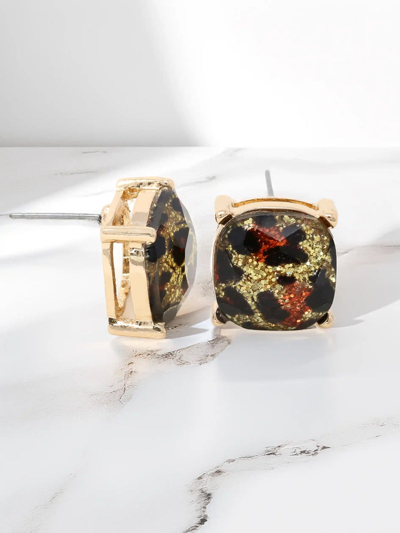 A pair of gold-tone stud earrings with cushion-cut black and gold marbled resin centers, displayed on a white marble surface.
