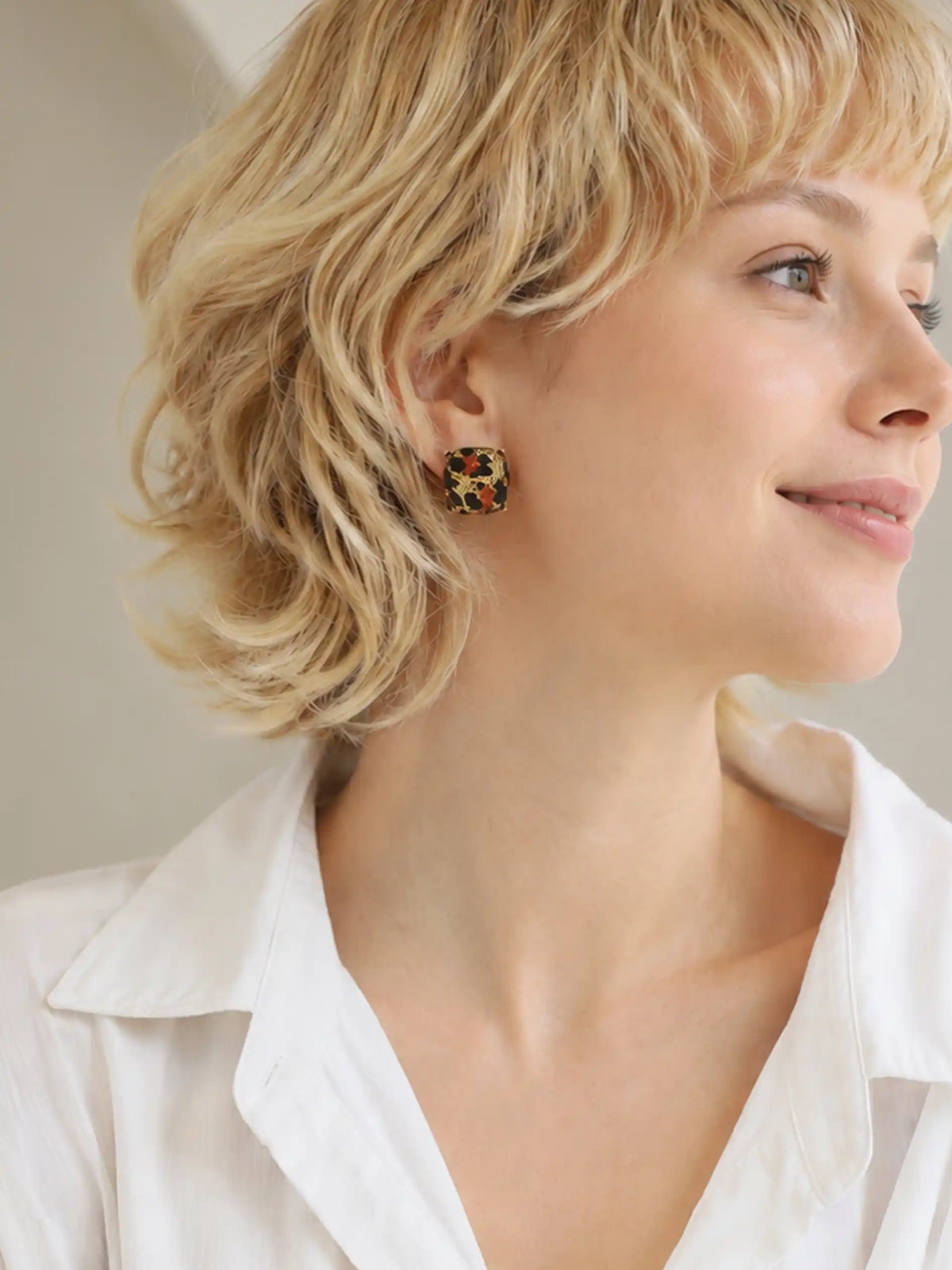 A close-up side profile of a woman with short blonde hair wearing a small square stud earring with a colorful leopard print pattern, paired with a white collared shirt.