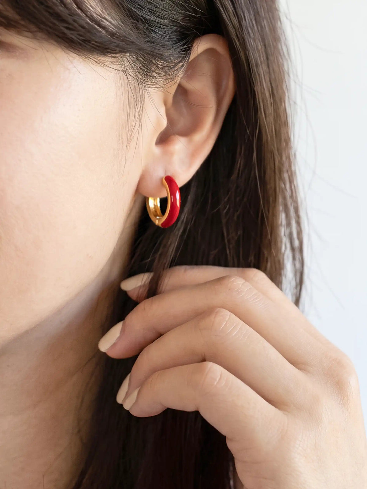 A close-up of a small gold-plated huggie hoop earrings featuring contrasting colorful red epoxy accents in a modern, compact design.