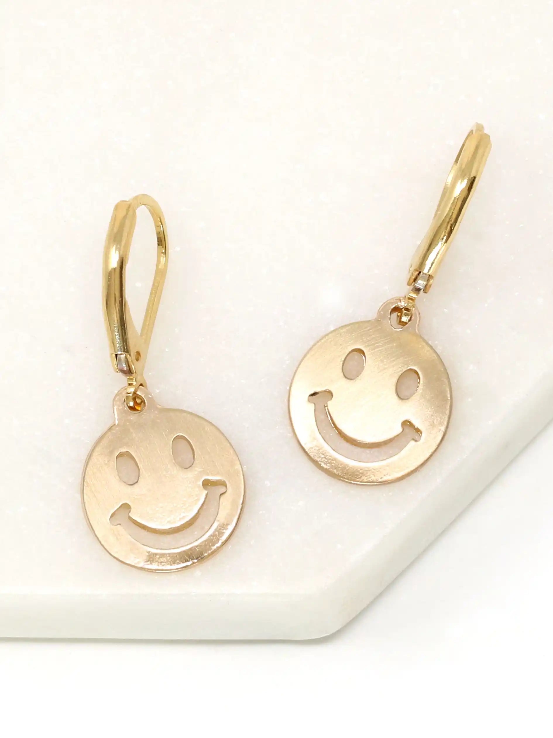 A pair of gold smiley face drop earrings with leverback hooks, displayed on a white textured surface.