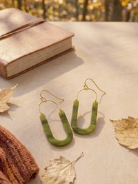 Olive green marbled U-shaped drop earrings with gold wire hooks, styled on a beige surface with autumn leaves, a brown knit sweater, and a book in the background.