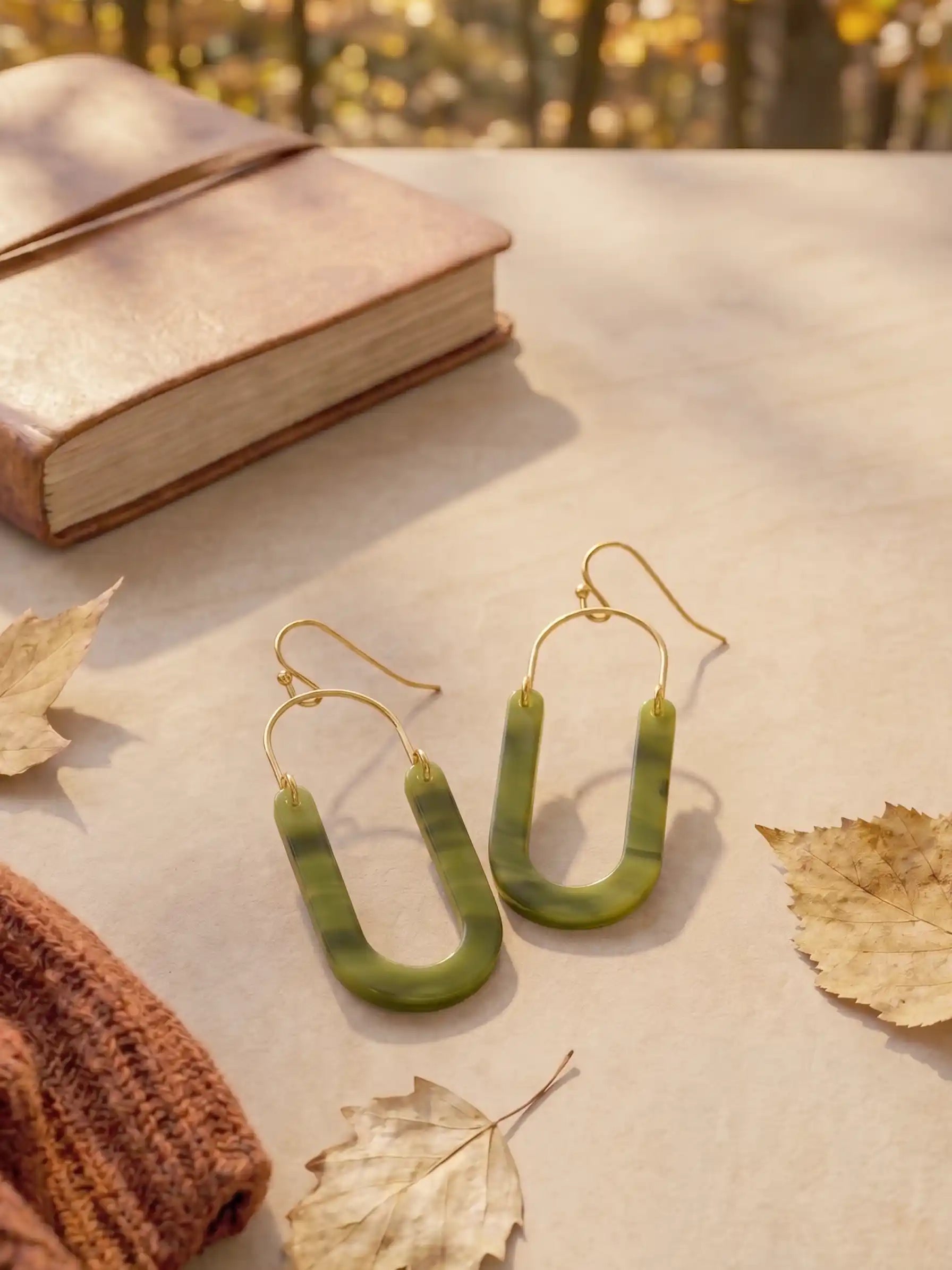 Olive green marbled U-shaped drop earrings with gold wire hooks, styled on a beige surface with autumn leaves, a brown knit sweater, and a book in the background.