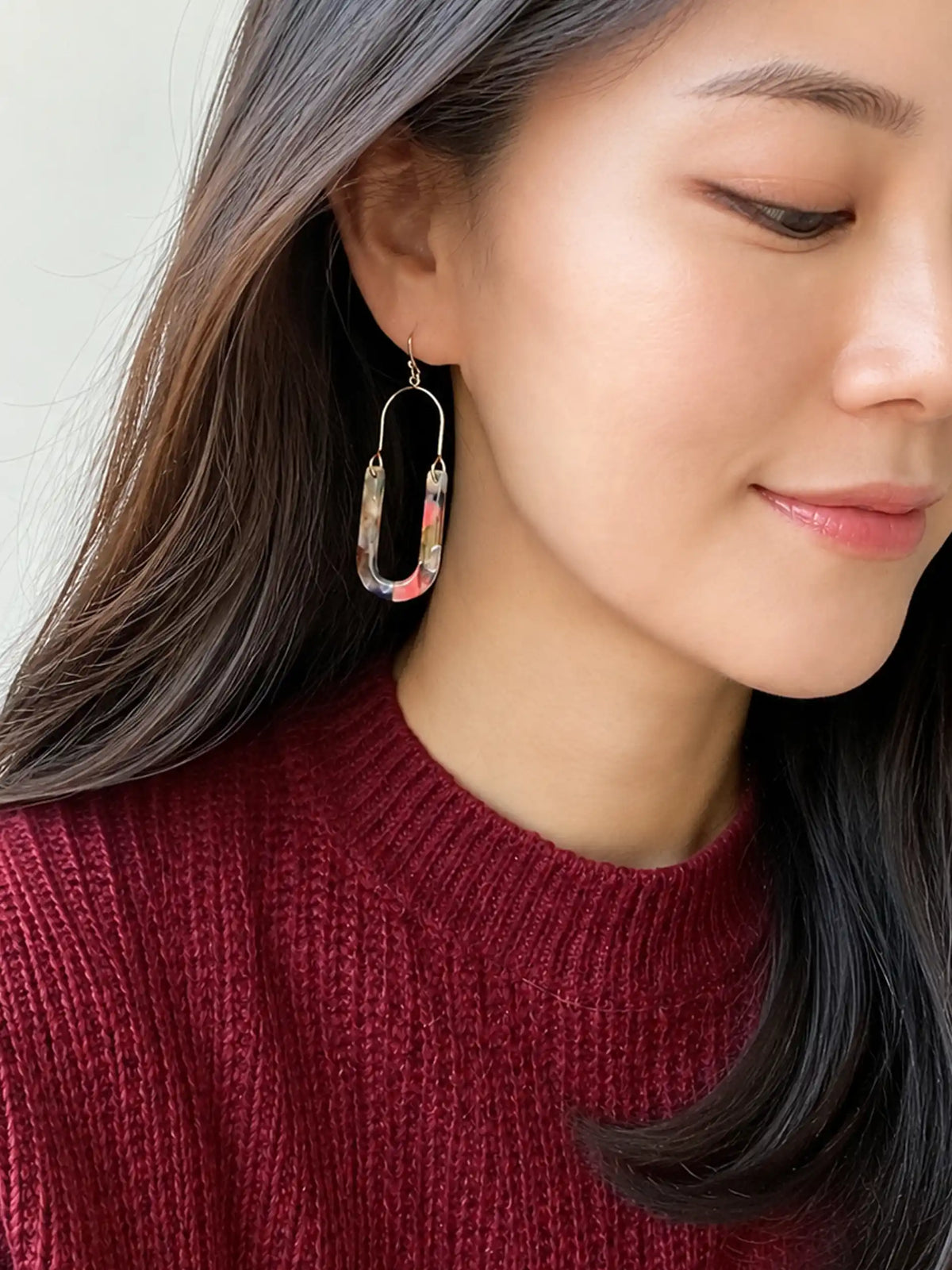 A close-up lifestyle shot of a woman wearing a large, U-shaped paperclip-style drop earring with a multicolor marbled pattern in pink, blue, and white, set in silver-toned metal.
