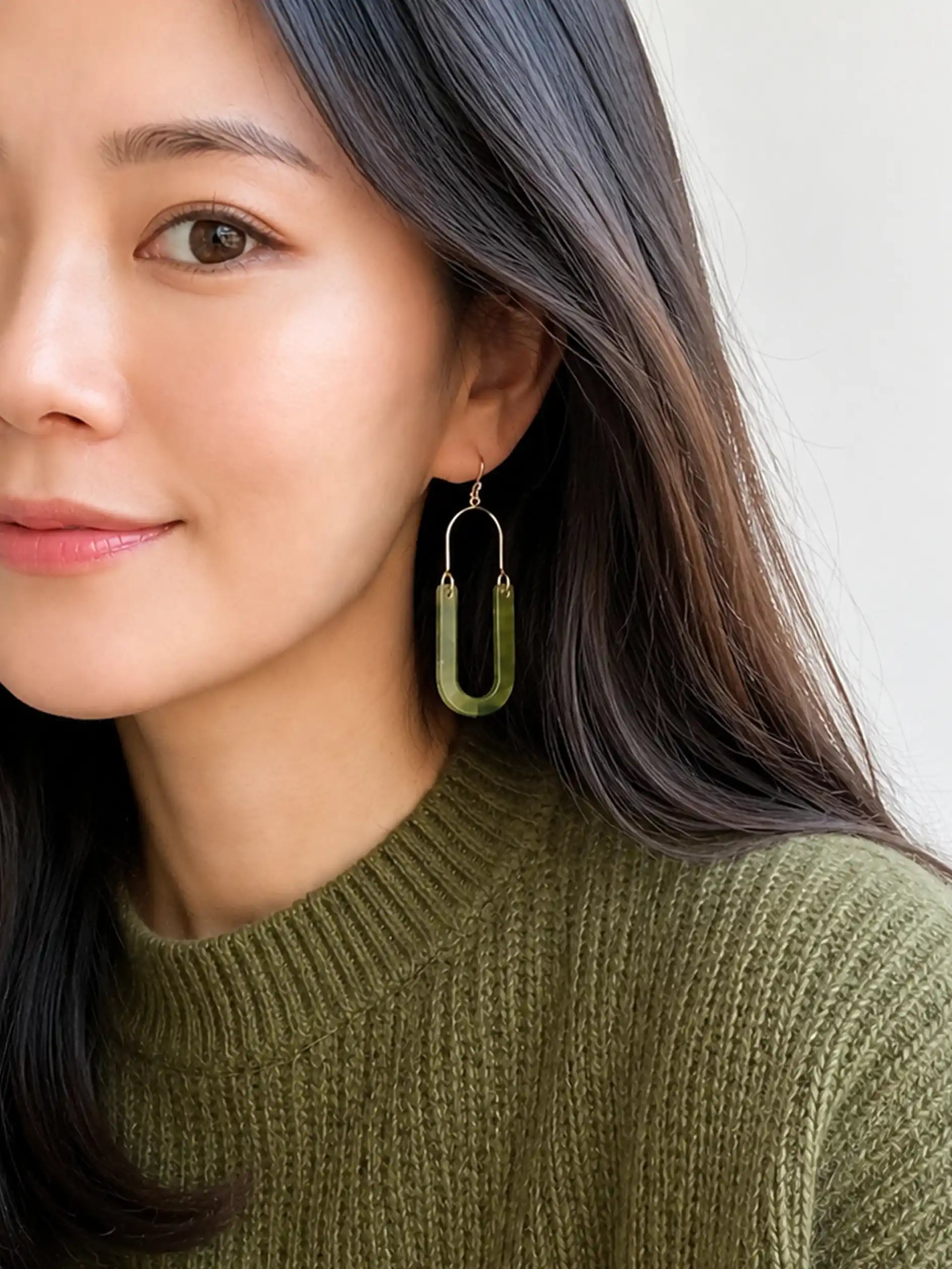 A close-up of a woman wearing a pair of green U-shaped drop earrings with silver-toned metal hooks and wires.