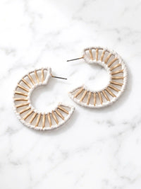 A pair of handmade crescent-shaped hoop earrings made from natural wood matchsticks wrapped in white raffia, displayed on a white marble surface.