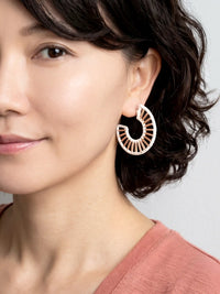 A close-up of a woman wearing a large, gold-toned crescent-shaped hoop earring with a sunburst or fan-like cutout pattern.