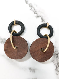 Alt Text: A pair of dangling earrings lying flat side-by-side on a white marble surface with grey veining. Each earring features a small, textured black ring at the top, which is connected to a larger, flat, brown circular wooden disc by a matte-gold metallic hoop that passes through the disc's center hole. A small gold-tone stud fixture loops over the top of each black ring.