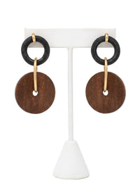 A pair of dangling earrings hanging side-by-side on a white T-bar jewelry display stand, shown against a bright white background. Each earring features a small, textured black ring at the top, which is connected to a larger, flat, brown circular wooden disc by a matte-gold metallic hoop that passes through the disc's center hole. A small gold-tone stud fixture loops over the top of each black ring
