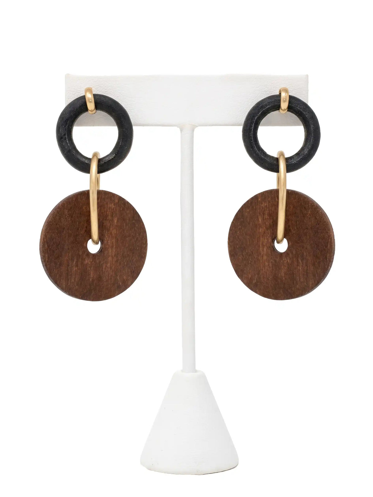 A pair of dangling earrings hanging side-by-side on a white T-bar jewelry display stand, shown against a bright white background. Each earring features a small, textured black ring at the top, which is connected to a larger, flat, brown circular wooden disc by a matte-gold metallic hoop that passes through the disc's center hole. A small gold-tone stud fixture loops over the top of each black ring