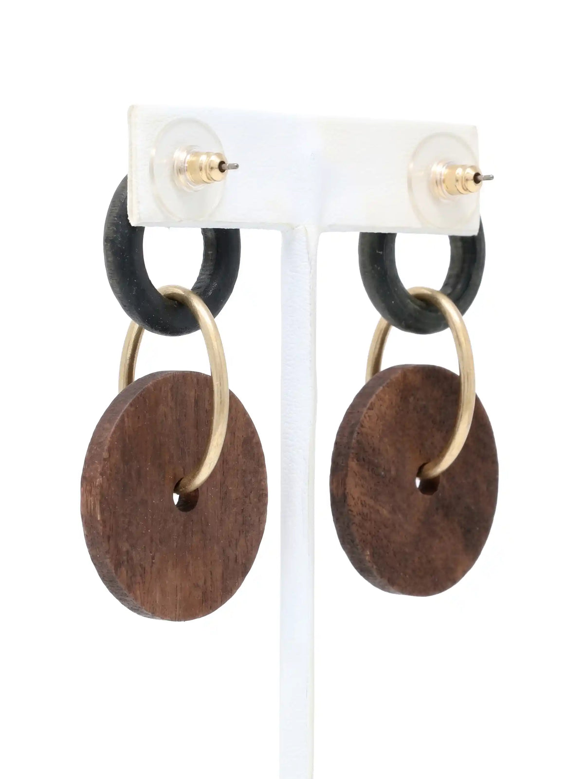 A pair of dangling earrings hanging side-by-side on a white T-bar jewelry display stand, shown against a bright white background. Each earring features a small, textured black ring at the top, which is connected to a larger, flat, brown circular wooden disc by a matte-gold metallic hoop that passes through the disc's center hole. A small gold-tone stud fixture loops over the top of each black ring