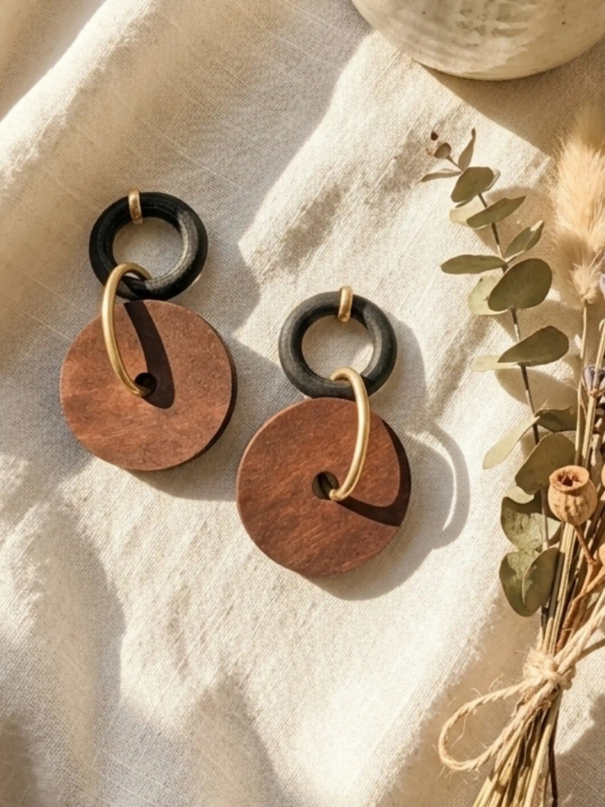 A pair of dangling earrings resting on a textured, cream-colored cloth bathed in warm sunlight, casting distinct shadows. Each earring features a small black ring at the top, connected to a larger, flat, brown circular wooden disc by a matte-gold metallic hoop that passes through the disc's center hole. A small gold-tone stud fixture loops over the top of each black ring. To the right of the earrings lies a small tied bundle of dried eucalyptus leaves and botanicals.