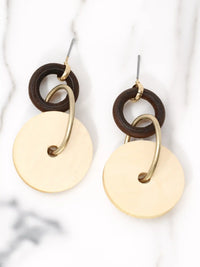 A pair of dangling earrings lying flat side-by-side on a white marble surface with grey veining. Each earring features a small dark brown wooden ring at the top, connected to a larger, flat, light beige circular wooden disc by a matte-gold metallic hoop that passes through the disc's center hole. A gold-tone stud post is attached to the top of each dark brown ring.