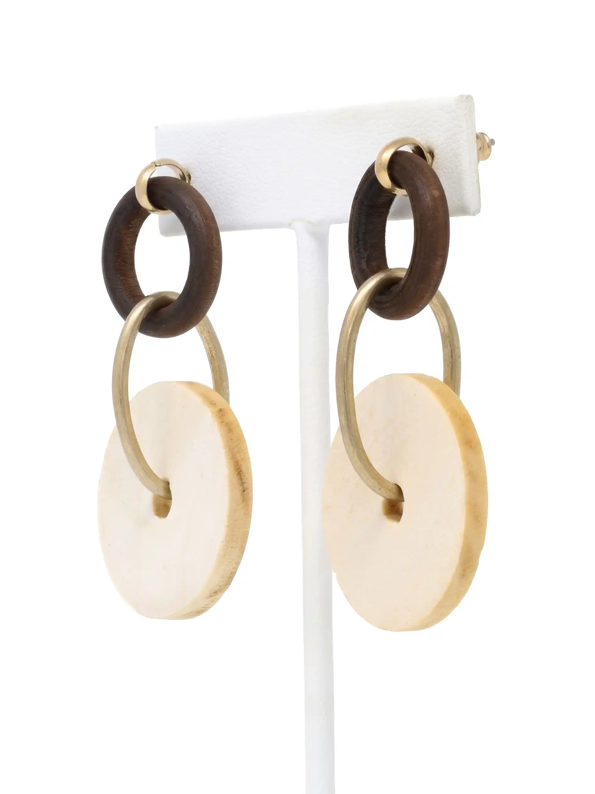 A pair of dangling earrings hanging side-by-side on a white T-bar jewelry display stand, shown against a bright white background. Each earring features a small dark brown, textured wooden ring at the top, which is connected to a larger, flat, light-beige circular drop disc made of natural wood by an elongated, matte-gold metallic hoop that passes through the center holes. The earrings are seen from the front, showcasing their earthy materials, natural grain, and modern, geometric design.