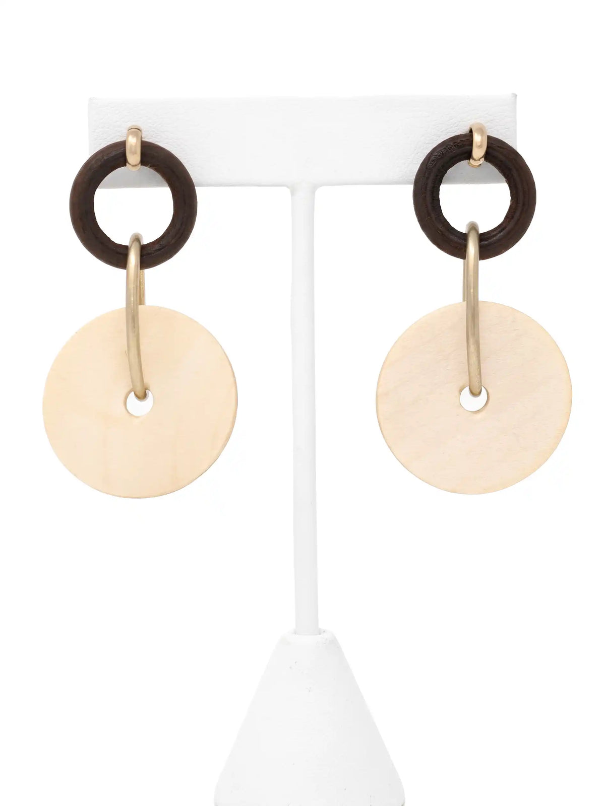 A pair of dangling earrings hanging side-by-side on a white T-bar jewelry display stand, shown against a bright white background. Each earring features a small dark brown, textured wooden ring at the top, which is connected to a larger, flat, light-beige circular drop disc made of natural wood by an elongated, matte-gold metallic hoop that passes through the center holes. The earrings are seen from the front, showcasing their earthy materials, natural grain, and modern, geometric design.