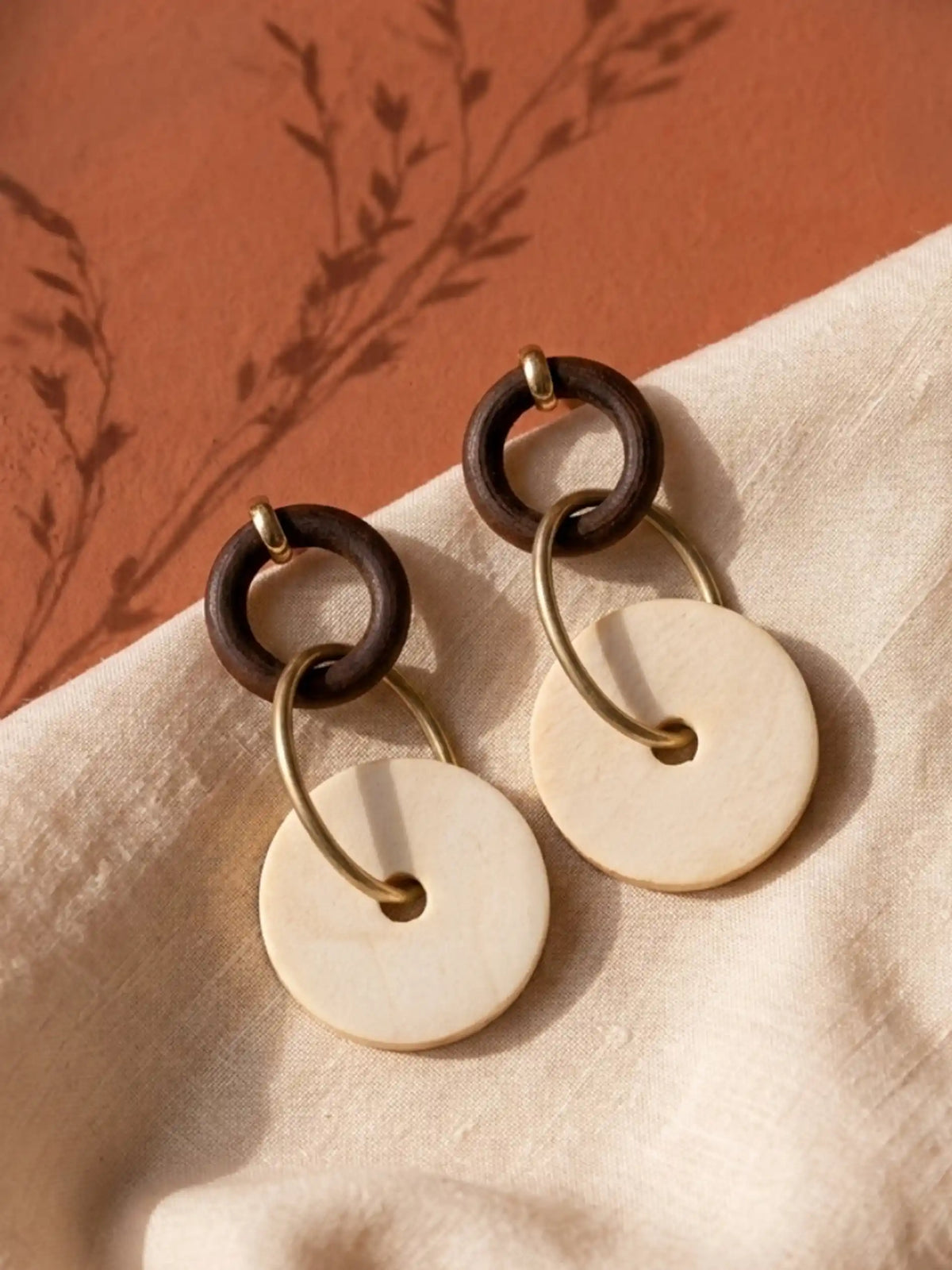A pair of dangling earrings resting on a textured, light beige fabric against a warm terracotta-colored background. Each earring features a small dark brown wooden ring at the top, connected to a larger, flat, light beige circular wooden disc by a matte-gold metallic hoop that passes through the disc's center hole. A small gold-tone stud fixture loops over the top of each dark brown ring. The scene is bathed in warm sunlight, casting delicate, botanical silhouettes as shadows across the surface.