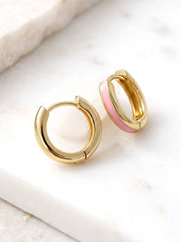 A close-up of a pair of 15mm gold-plated huggie hoop earrings with a light pink epoxy color accent on one side.