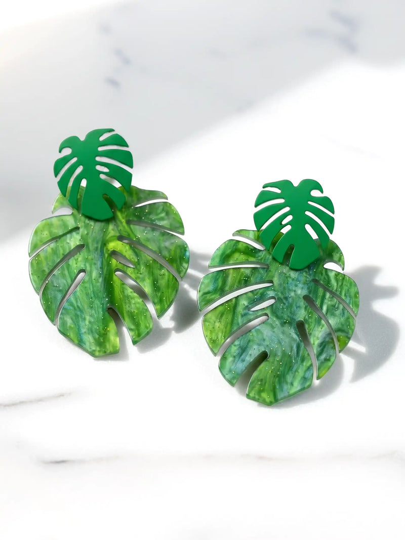 A pair of green monstera leaf stud earrings with a glossy marbled green resin finish, displayed on a light gray marble surface.