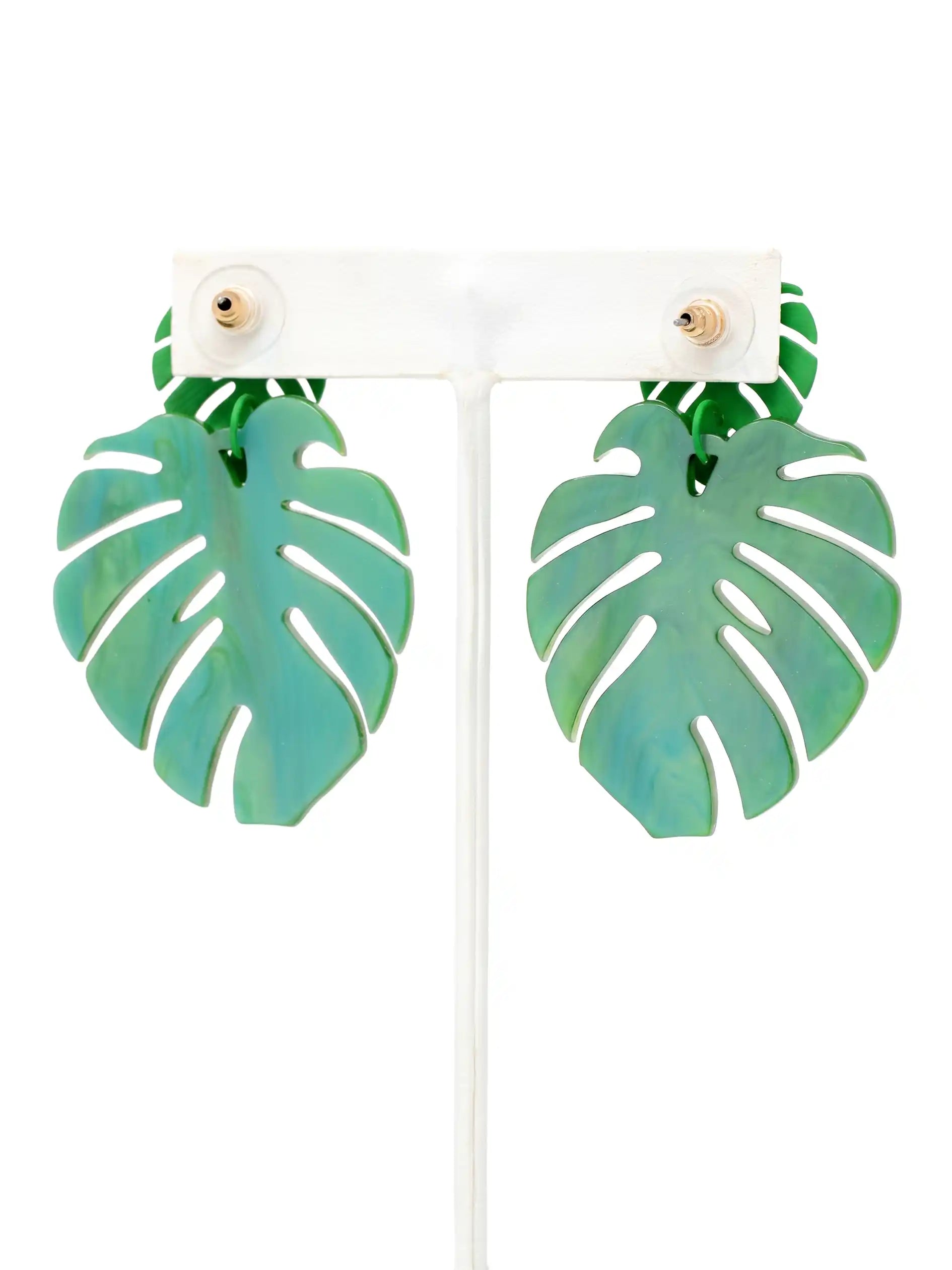 A pair of green monstera leaf drop earrings displayed on a white T-shaped jewelry stand against a white. background.