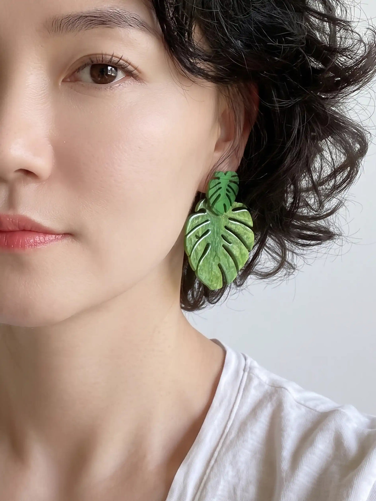 A close-up of a woman wearing a green monstera leaf-shaped drop earring, showing the earring against her ear and dark hair.