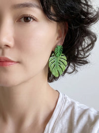 A close-up of a woman wearing a green monstera leaf-shaped drop earring, showing the earring against her ear and dark hair.