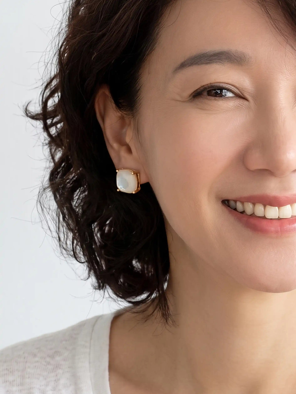 A close-up of a woman wearing a gold-toned stud earring with a white rectangular cabochon stone, set against a clean white background.