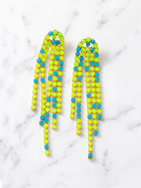 Overhead shot of a pair of long, cascading duster earrings featuring tiered strands of vibrant neon yellow-green and bright turquoise-blue faceted beads set in gold-tone metal. The earrings are lying flat on a white marble surface.
