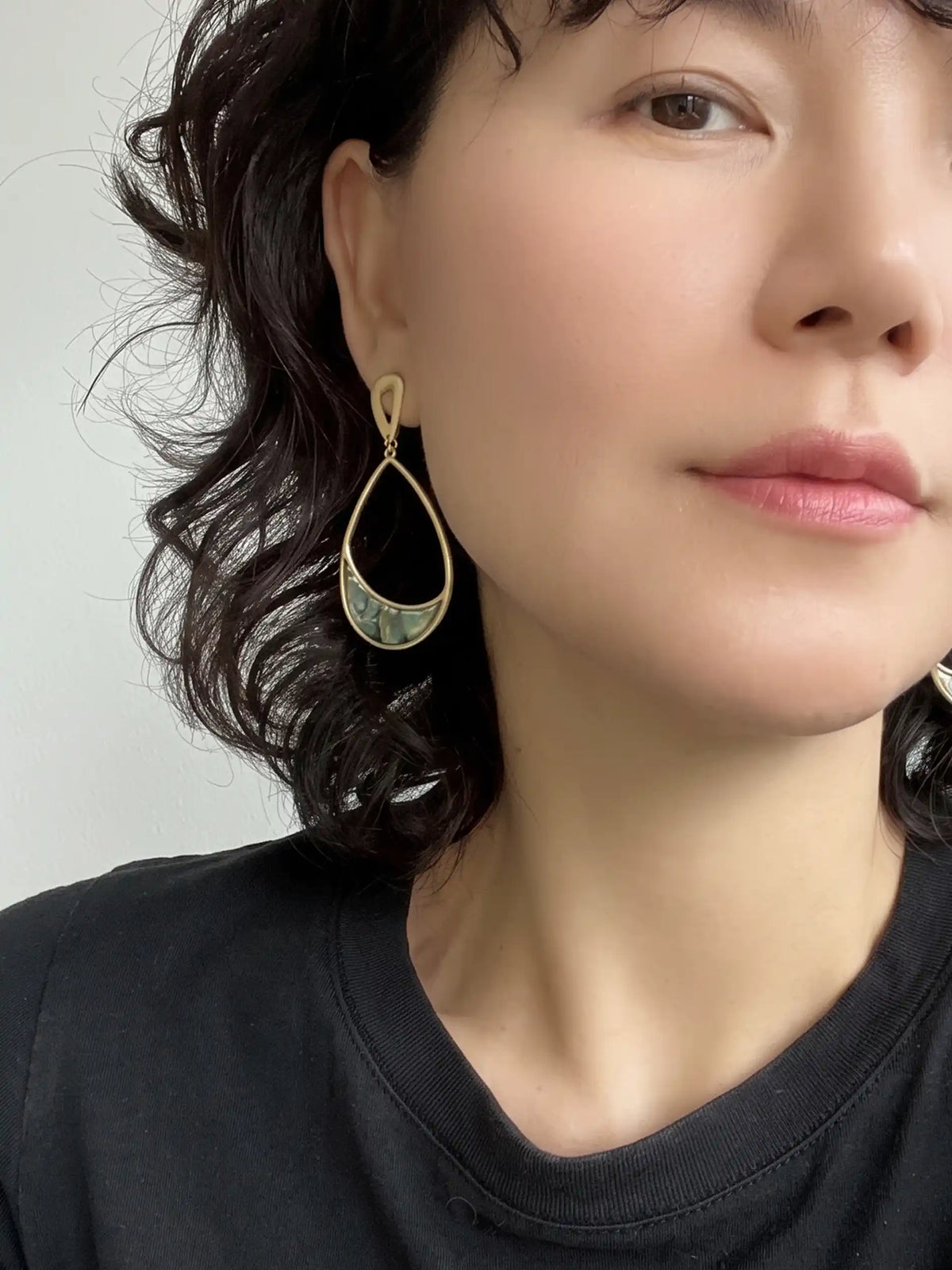 A close-up of a woman wearing a gold teardrop-shaped dangle earring with a green and blue patterned center, paired with a black crewneck shirt.