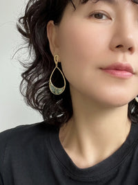 A close-up of a woman wearing a gold teardrop-shaped dangle earring with a green and blue patterned center, paired with a black crewneck shirt.
