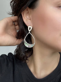 A close-up of a person wearing a dangling teardrop earring with a silver-tone frame and a green iridescent inlay, shown against skin and dark hair.