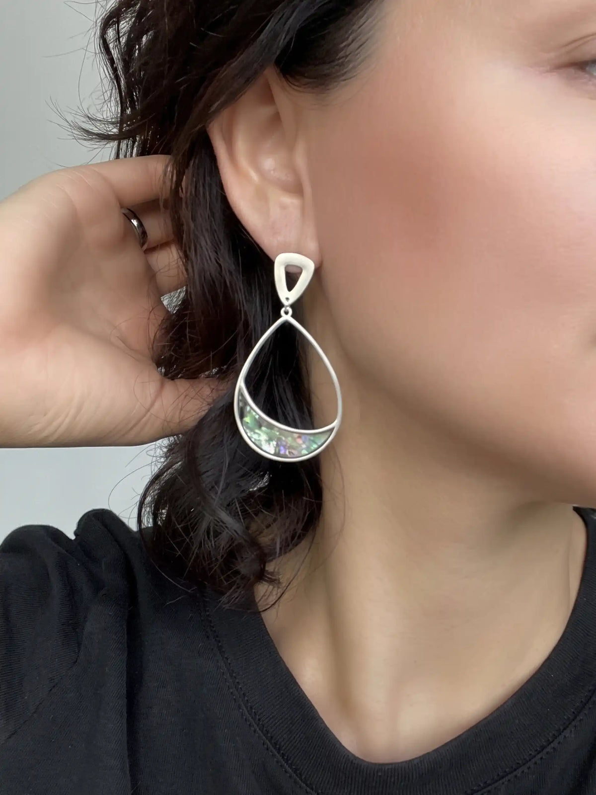 A close-up of a person wearing a dangling teardrop earring with a silver-tone frame and a green iridescent inlay, shown against skin and dark hair.