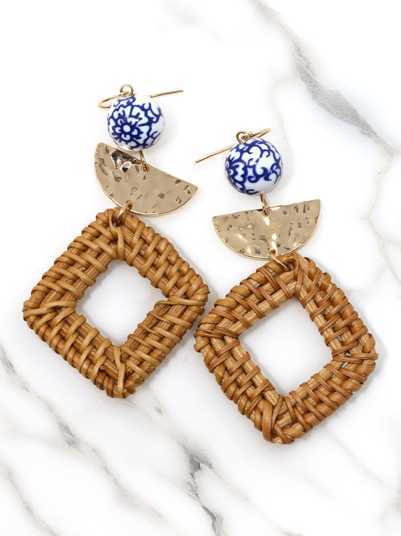 Pair of earrings with woven brown design, blue and white spherical beads, and gold accents on a marble surface.