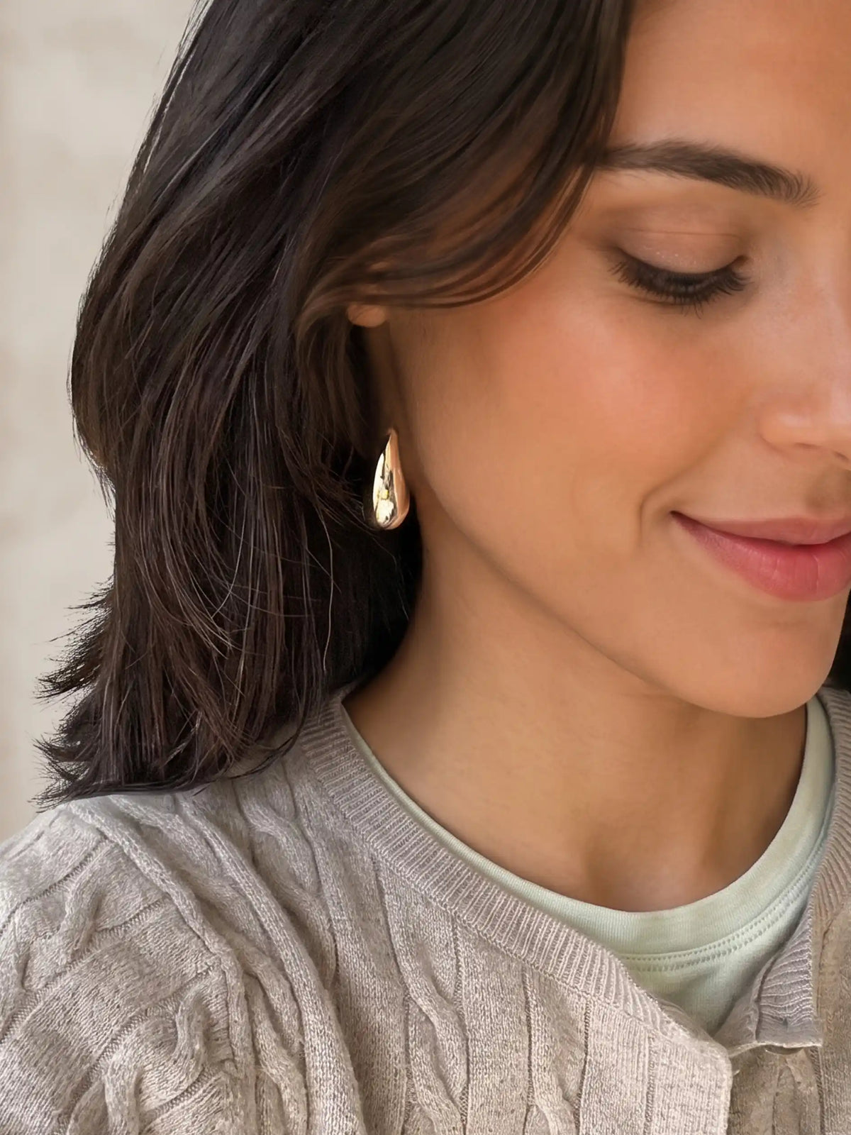 A close-up of a woman wearing a small gold teardrop-shaped dangle earring, paired with a light sage green crewneck shirt and a textured beige cardigan.