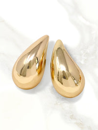 A pair of teardrop-shaped gold earrings with a highly polished, reflective metallic finish, displayed on a light marble surface.