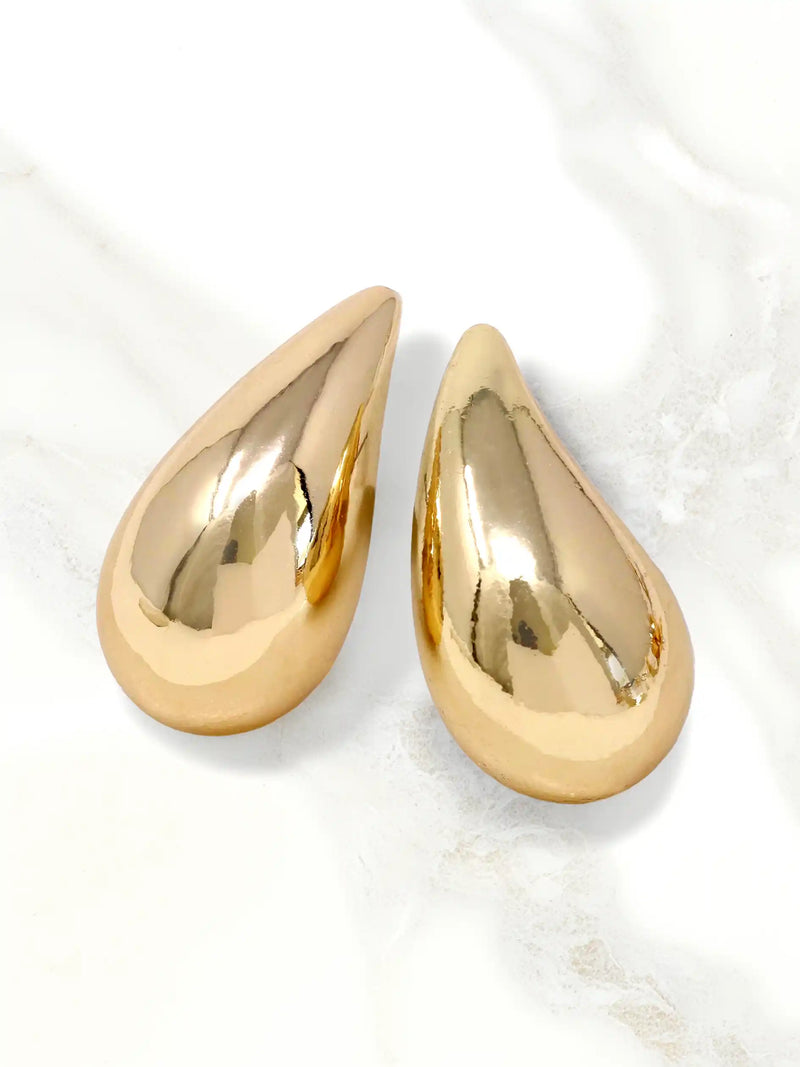 A pair of teardrop-shaped gold earrings with a highly polished, reflective metallic finish, displayed on a light marble surface.