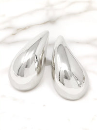 A pair of polished silver teardrop-shaped earrings with a highly reflective, metallic finish, displayed on a white marble surface.
