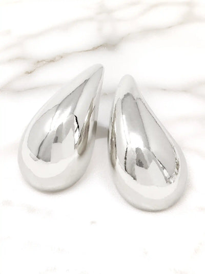 A pair of polished silver teardrop-shaped earrings with a highly reflective, metallic finish, displayed on a white marble surface.