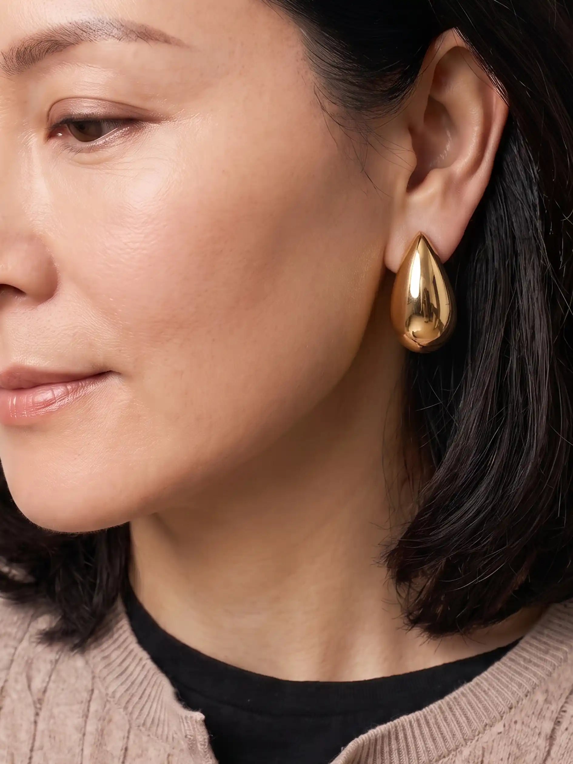 A close-up of a woman wearing a polished gold teardrop-shaped drop earring, showing the side profile of her face and ear.