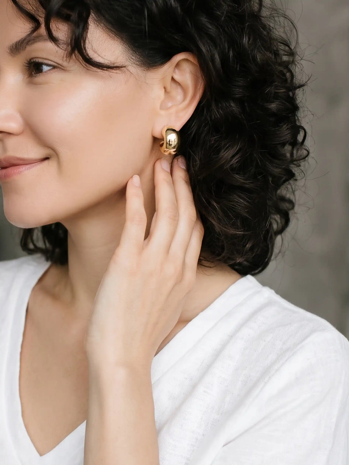 A close-up lifestyle shot of a woman with dark curly hair wearing a small gold hoop earring, touching her ear with her hand. She is wearing a white V-neck top against a soft gray background.