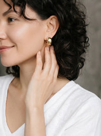 A close-up lifestyle shot of a woman with dark curly hair wearing a small gold hoop earring, touching her ear with her hand. She is wearing a white V-neck top against a soft gray background.