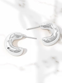 A pair of polished silver crescent-shaped hoop earrings with post backs, displayed on a white marble surface.