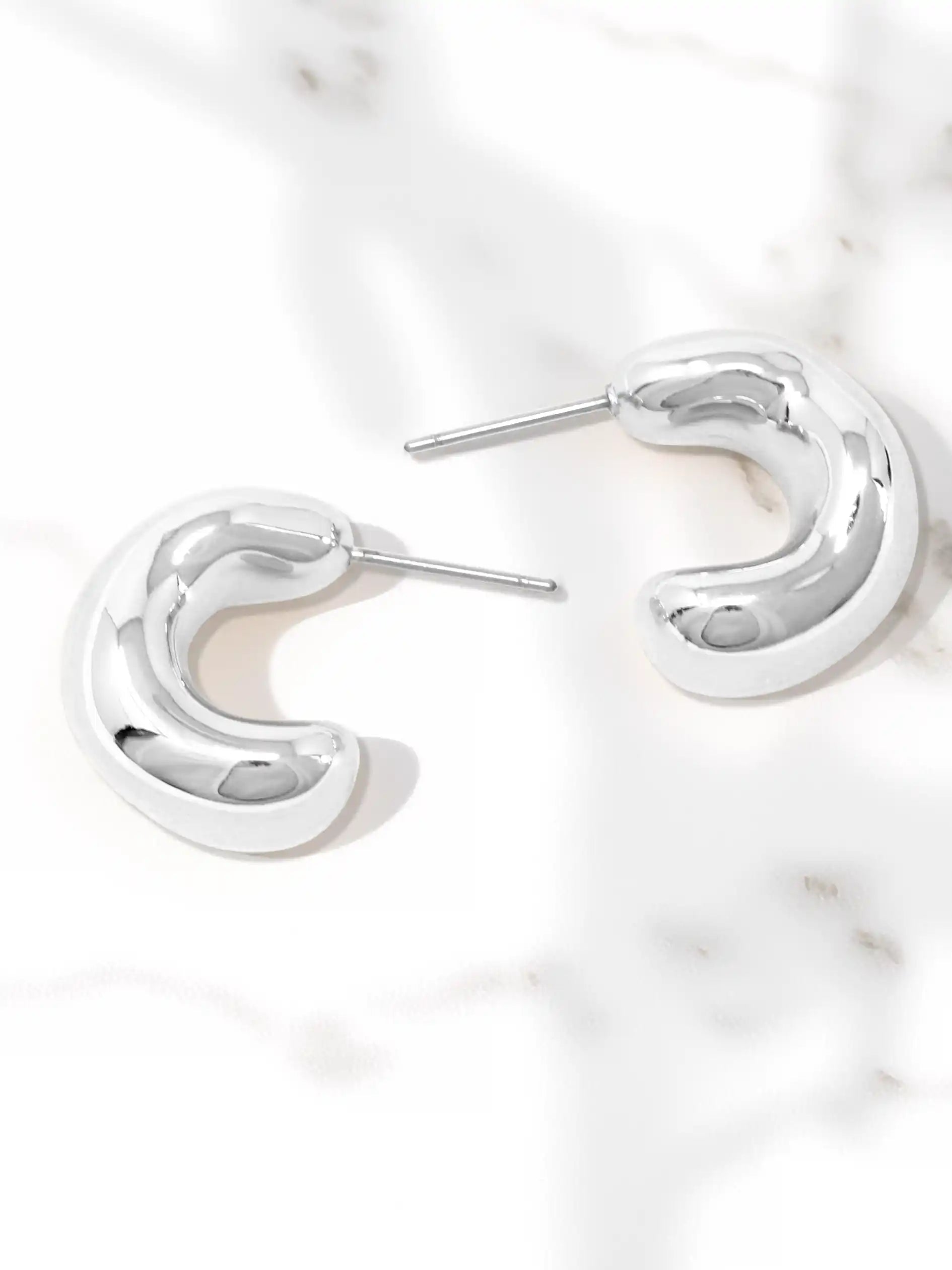 A pair of polished silver crescent-shaped hoop earrings with post backs, displayed on a white marble surface.
