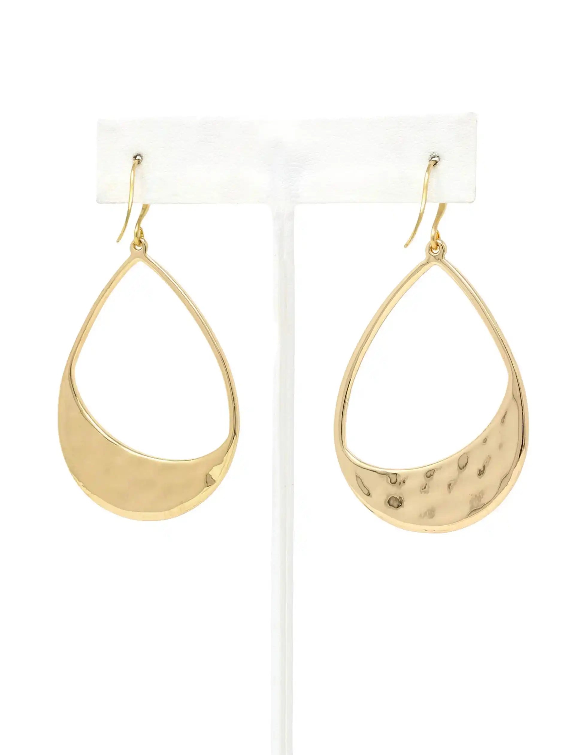 A pair of gold teardrop-shaped dangle earrings with one smooth curved edge and one hammered textured edge, displayed on a white plastic jewelry stand against a white background.