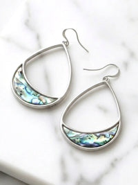 A pair of silver teardrop dangle earrings with a crescent-shaped inlay of iridescent abalone shell, displayed on a white marble surface.