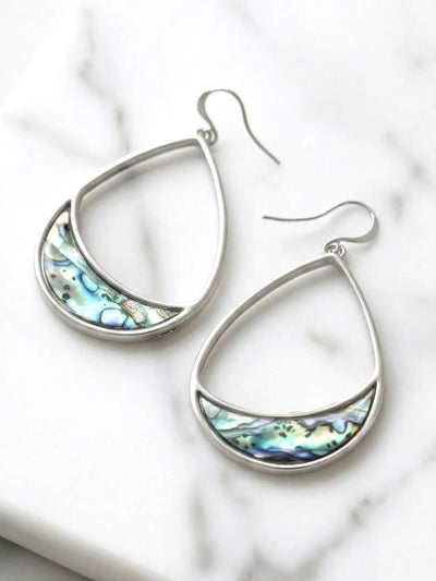 A pair of silver teardrop dangle earrings with a crescent-shaped inlay of iridescent abalone shell, displayed on a white marble surface.