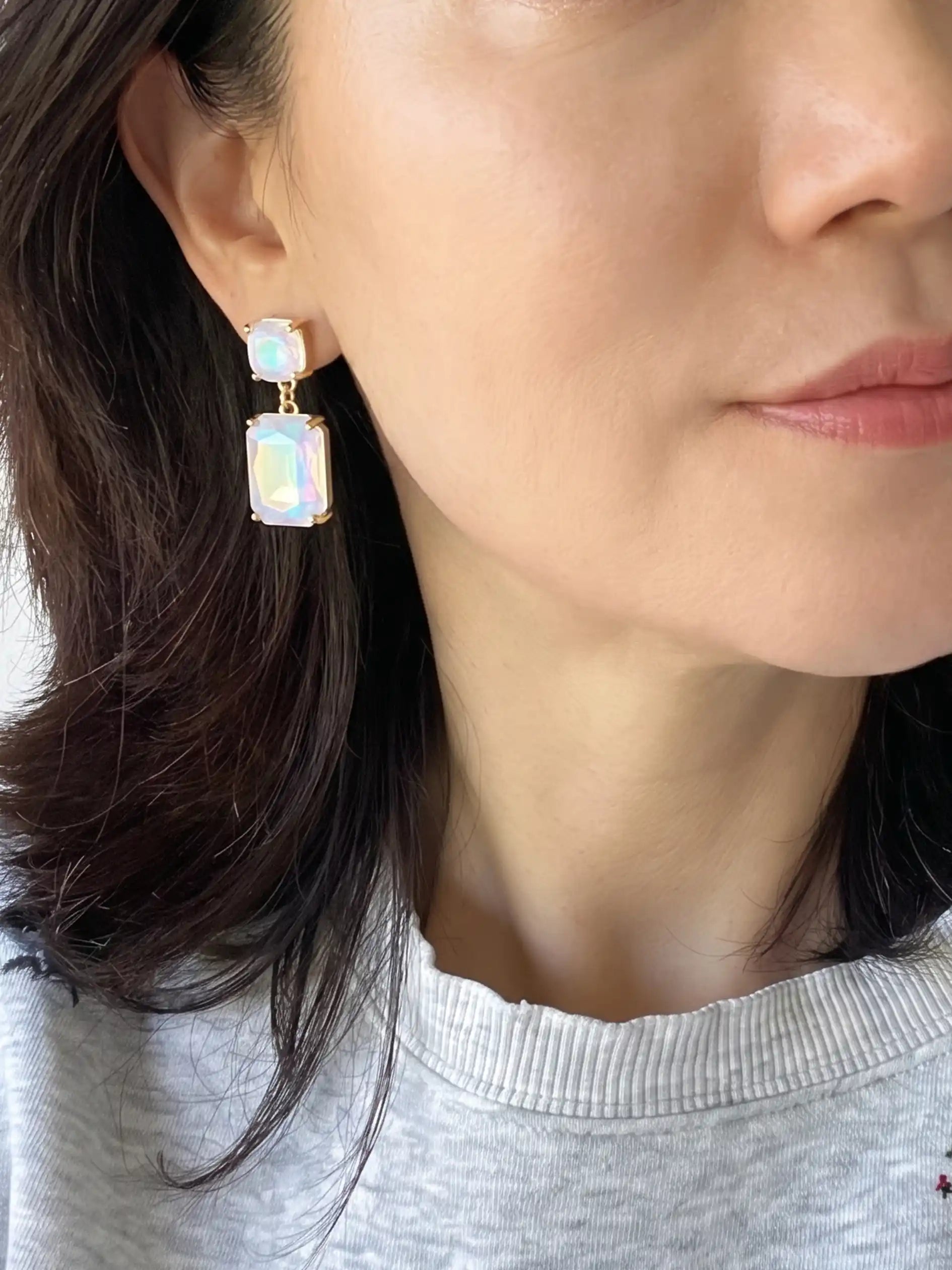 A close-up of a woman wearing a gold drop earring with two iridescent, color-shifting rectangular gemstones.