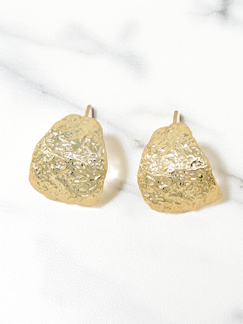 A pair of gold-tone textured bold hoop earrings with an organic, hammered finish, displayed on a marble surface.