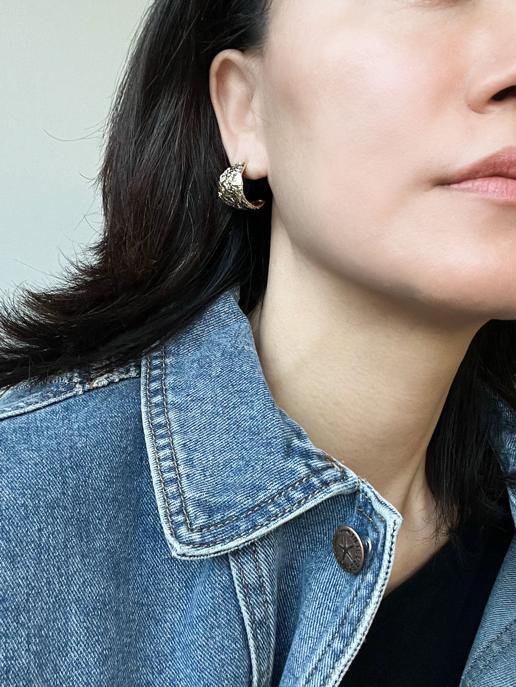 A pair of gold-tone textured bold hoop earrings with an organic, hammered finish, displayed on a marble surface.