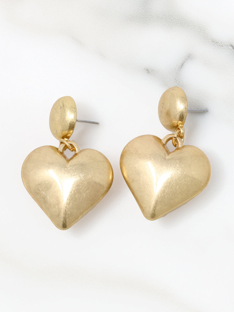 A pair of gold-tone puffy heart statement earrings featuring a smooth, polished finish. Each earring has a rounded stud at the top with a dangling puffed heart charm, creating a bold yet elegant look. Perfect for adding a touch of glamour to any outfit.